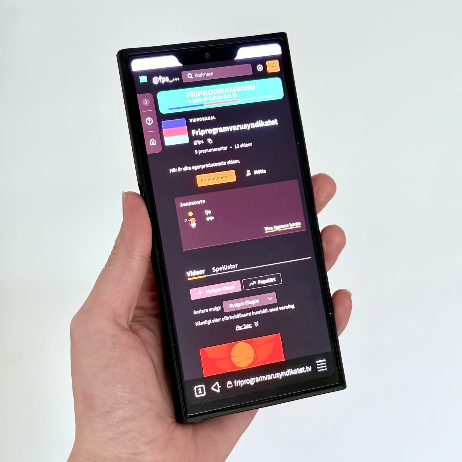 Miranda holding the Jolla Phone 2026 in left hand. Showing Friprogramvarusyndikatets Peertube platform with channel for our videos in Gecko ESR v115 web browser.